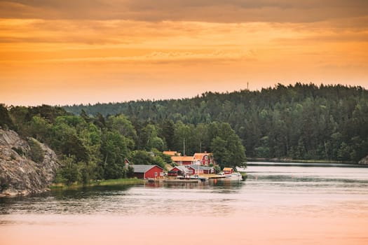 Sweden. Many Beautiful Red Swedish Wooden Log Cabins Houses On Rocky Island Coast In Summer Evening. Lake Or River Landscape.
