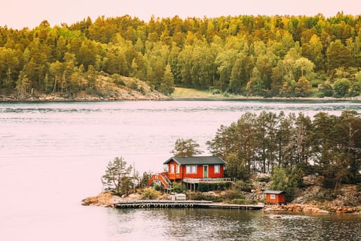 Sweden. Many Beautiful Red Swedish Wooden Log Cabin House On Rocky Island Coast In Summer. Lake Or River Landscape.