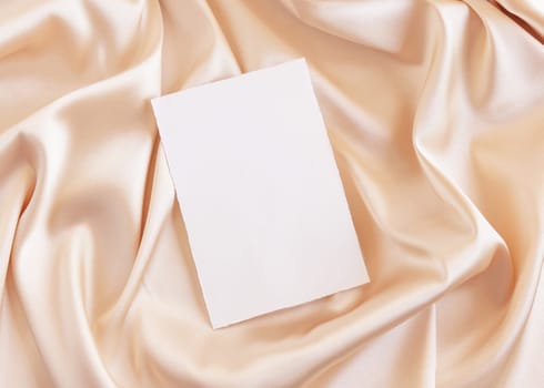 Blank card on beige silky fabric drapery top view, mockup. Romantic scene with vertical paper card, greeting or wedding Invitation. .