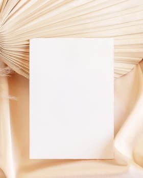 Blank card on beige satin fabric and dried palm leaf close up, mockup. Boho scene with vertical paper card. Bohemian or close to nature greeting or wedding Invitation. Romantic composition.
