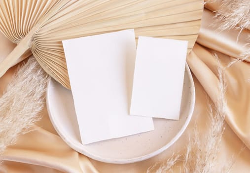 Blank cards on plate with dried palm leaf and beige silky fabric close up, mockup. Romantic scene with vertical paper cards. Bohemian pastel greeting or wedding Invitation .