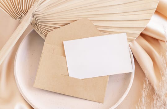 Blank card and envelope on plate with dried palm leaf and beige silky fabric close up, mockup. Romantic scene with horizontal paper card. Pastel greeting or wedding stationery.