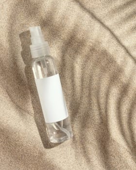 Cosmetic spray bottle with blank label on beige sand top view. Packaging mockup. Natural skincare product for beauty routine. Summer sunny day with palm leaf hard shadow, tropical vacation mood