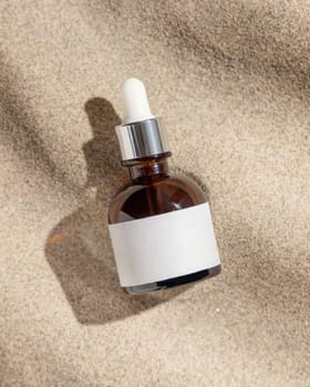 Dropper bottle with blank label on beige sand top view, hard light. Package mockup. Natural skincare product. Facial cosmetics for beauty routine. Zen style with waves on sand, minimal aesthetic
