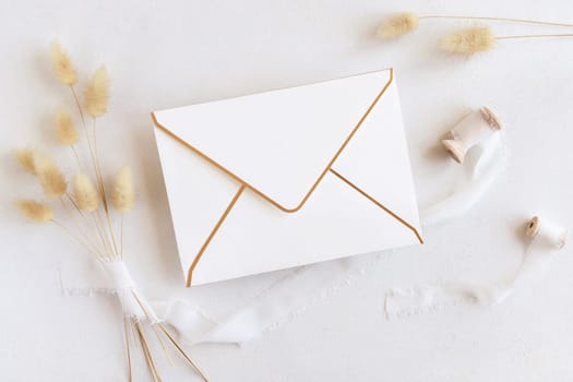 Blank envelope near silk ribbons and dried hare's tail grass top view on white, mockup. Boho romantic scene. Bohemian or close to nature greeting or wedding Invitation