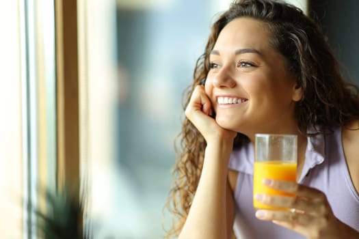 Happy woman holding orange juice looking through window at home