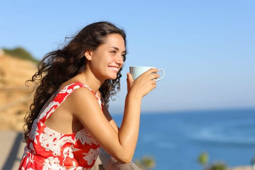 Happy tourist drinking coffee looking away on the beach on summer vacation