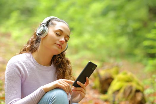 Beautiful woman sitting in a forest listening to music with headphones and phone relaxing