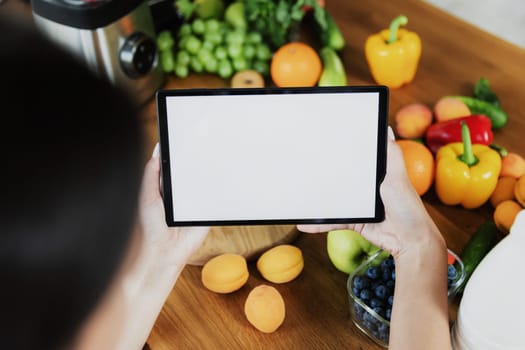 Female hands holding tablet with empty blank white screen mockup in front of vegetables, fruits on table in kitchen at home. Selective focus, cropped. Food delivery concept, mobile diet plan app