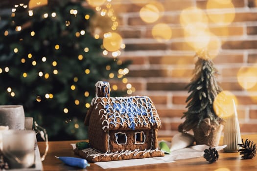 Little homemade gingerbread house standing on table with decorations, candles in living room decorated with defocused New Year lights, Christmas fir tree. Wonderful happy holiday mood. Copy space
