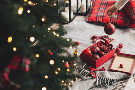 Little child girl decorates Christmas fir tree with red ornament balls. Small kid holding gift in hand before hanging it to tree with light, garland. Box with balls laying on floor with decorations
