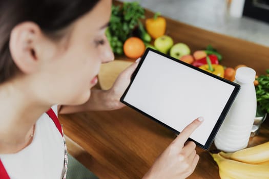 Young woman using smartphone, girl hands holding and clicking on tablet with empty blank white screen mockup. Fresh food on table at kitchen. Cooking healthy weight loss menu. Mobile diet plan app