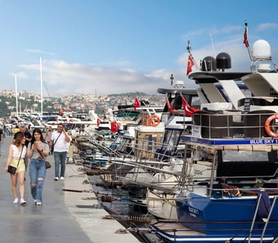 Istanbul, Turkey - August 31, 2022: People walking on side walk by the Bosphorus Strait between Bebek and Hisari districts, beside docked motor boats and yachts, against cloudy blue sky on sunny day