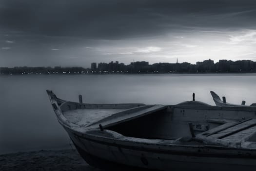 Black and white shot of fishing boat on the beach at sunrise with Egyptian Alexandria city skyline in far distance and cloudysky at sunrise