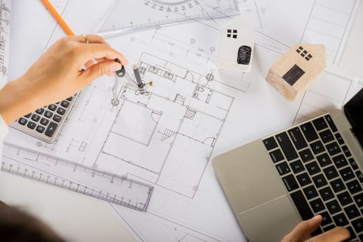Architectural project workplace. Architect typing laptop keyboard to review design of house before editing or sketching with compass on plan blueprint paper on table desk at architecture office