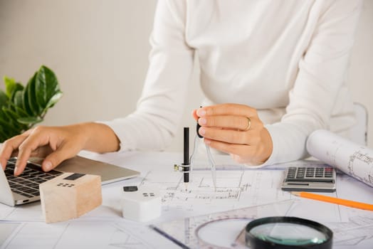 Architectural project workplace. Architect typing laptop keyboard to review design of house before editing or sketching with compass on plan blueprint paper on table desk at architecture office