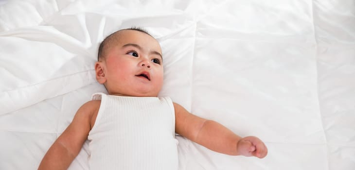 Asian smiling infant child at home, Cute little baby on bed under soft blanket, baby wearing bodysuit lying on white beedsheets in bedroom