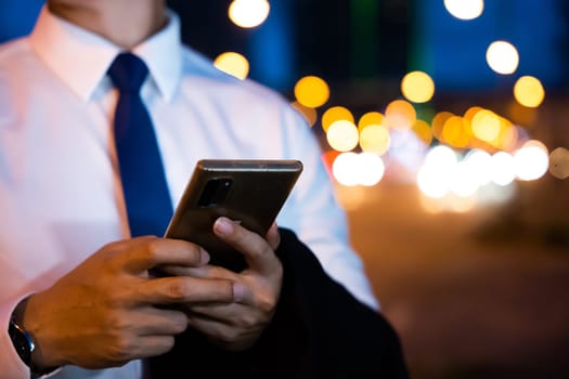 Portrait Asian businessman typing an sms message via smartphone after work near office at night city street, bokeh lights, young man walking and chatting with friends at social networks outdoor