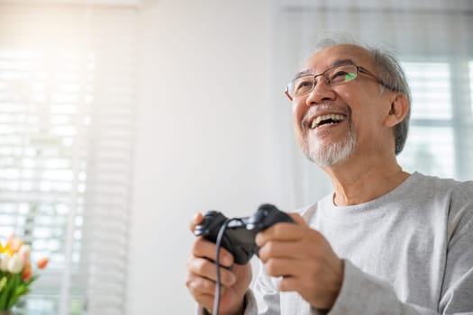 Asian senior old man enjoying holding joystick playing video game at home in living room, mature man hands using game controller, Funny on retirement elderly smile sitting on sofa life gaming
