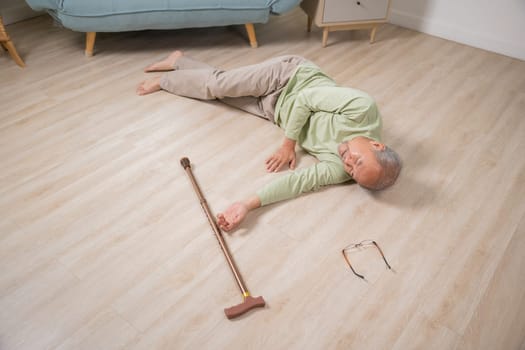 Asian senior man falling down lying on floor at home alone with wooden walking stick in living room, Elderly man headache after fall down, Health care and medicine concept