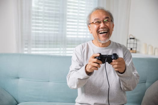 Asian senior old man enjoying holding joystick playing video game at home in living room, mature man hands using game controller, Funny on retirement elderly smile sitting on sofa life gaming