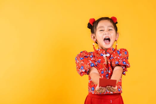 Chinese New Year. Happy Asian Chinese little girl smile wear red cheongsam handed angpao or red packet monetary gift, Portrait children in traditional dress, studio short isolated on yellow background