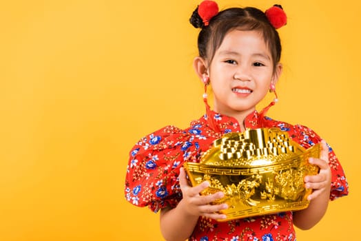 Chinese New Year. Happy Asian Chinese little girl smile wearing red cheongsam holding big gold ingot, Portrait children in traditional dress hold golden bar, studio short isolated on yellow background