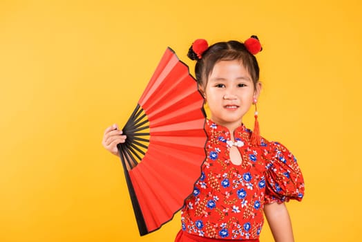 Chinese New Year. Happy Asian Chinese little girl smile wearing red cheongsam holding fan, Portrait children in traditional dress, studio short isolated on yellow background
