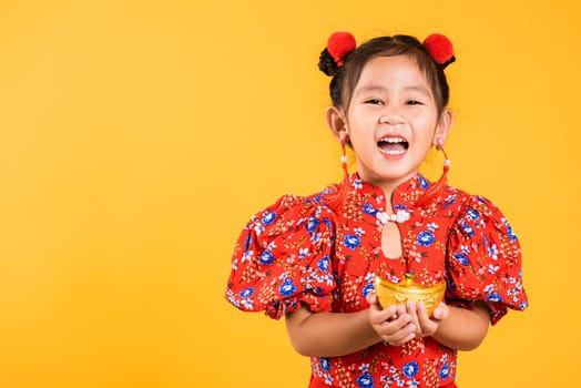 Chinese New Year. Happy Asian Chinese little girl smile wearing red cheongsam holding gold ingot, Portrait children in traditional dress hold golden bar, studio short isolated on yellow background
