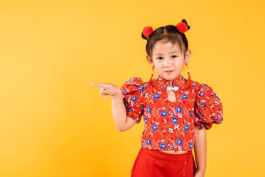 Happy Asian Chinese little girl wear red cheongsam qipao pointing finger to space, Portrait children in traditional dress point to empty, Chinese New Year, studio short isolated on yellow background