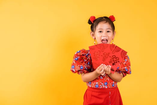 Chinese New Year. Happy Asian Chinese little girl smile wear red cheongsam qipao hold angpao red packet monetary gift, Portrait child in traditional dress, studio short isolated on yellow background