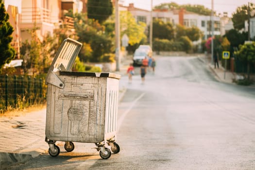 Metal Waste Container On Wheels In Street During Summer Sunny Evening. Container For Temporarily Storing Waste. Dustbin, Garbage Can, Trash Can.
