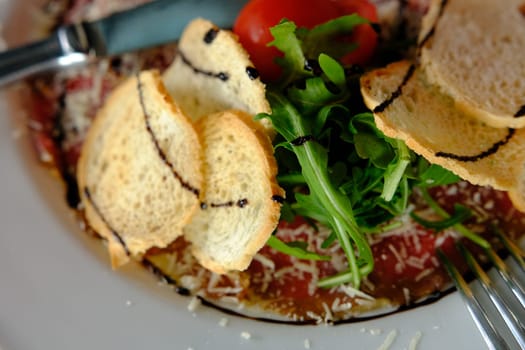 Beef Carpaccio cold appetizer with parmesan, tomatos and arugula on white plate with hardtack on dark background.