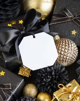 Black and golden Christmas Gift Boxes with a bow and ornaments close up, square paper gift tag mockup, copy space. Dark winter composition with blank label card for Christmas, New Year, Birthday, Anniversary