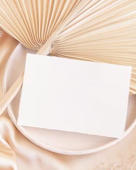 Blank card on plate with dried palm leaf and beige silky fabric close up, mockup. Romantic scene with horizontal paper card. Bohemian pastel greeting or wedding Invitation .