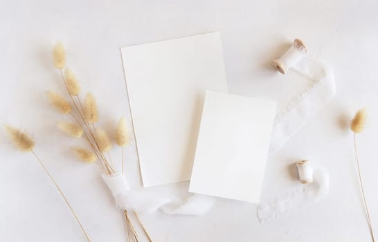 Blank cards near silk ribbons and dried hare's tail grass top view on white, mockup. Boho romantic scene with vertical cards. Bohemian or close to nature greeting or wedding Invitation