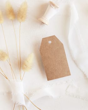 Blank gift tag near silk ribbons and dried hare's tail grass top view on white, mockup. Boho romantic scene with kraft paper gift tag. Bohemian or close to nature greeting