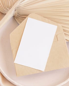 Blank card and envelope on plate with dried palm leaf and beige silky fabric close up, mockup. Romantic scene with vertical paper card. Pastel greeting or wedding stationery.