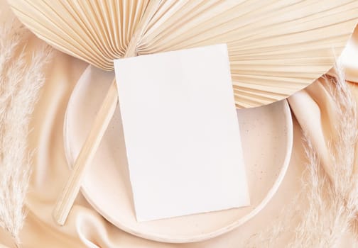 Blank card on plate with dried palm leaf and beige silky fabric close up, mockup. Romantic scene with vertical paper card. Bohemian pastel greeting or wedding Invitation .