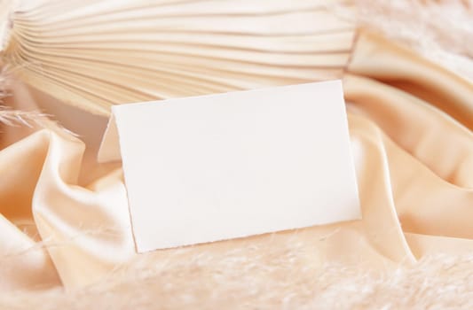 Folded place card on beige satin fabric and dried palm leaf close up, mockup. Boho scene with horizontal paper card. Bohemian or close to nature greeting or wedding Invitation. Romantic composition.