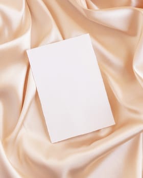 Blank card on beige silky fabric drapery top view, mockup. Romantic scene with vertical paper card, greeting or wedding Invitation. .