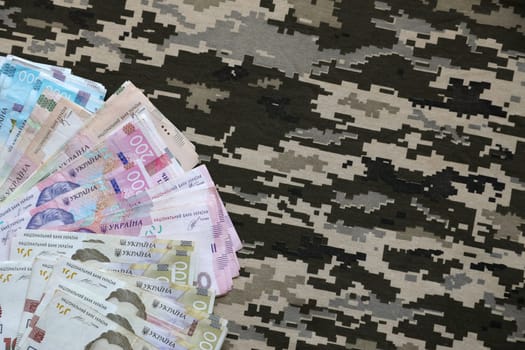 Ukrainian hryvnya bills on fabric with texture of Ukrainian military pixeled camouflage. Cloth with camo pattern in grey, brown and green pixel shapes. Official uniform of Ukrainian soldiers close up