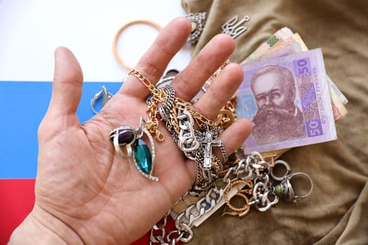 Marauders hand with bunch of stolen jewelry and money on military uniform cloth fabric and Russian flag. Looting by Russian soldiers in the Ukrainian cities during the Russian invasion