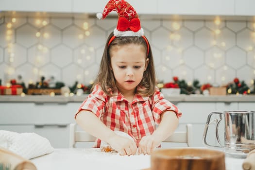 Little dark-haired girl 3 years old in red Christmas cap and checkered shirt prepares dough for gingerbread cookies in white Christmas decorated kitchen with garland lights. Merry Christmas, New Year