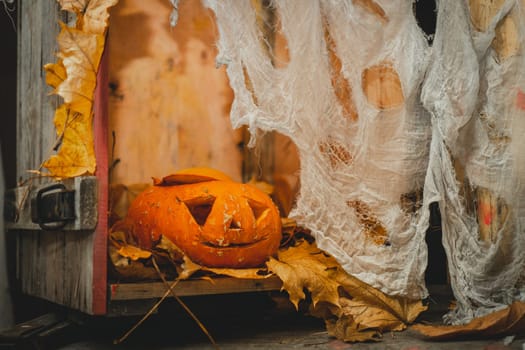 Orange bright halloween pumpkin with carved nose, mouth and eyes on wooden box with gauze as web next to autumn yellow leaves. Scary face carved on vegetable. Halloween holiday decoration