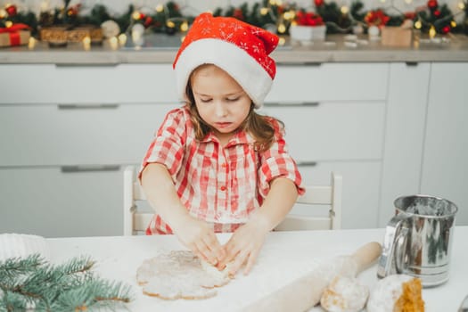 Little dark-haired girl 3 years old in red Christmas cap and checkered shirt cuts out gingerbread cookies from rolled dough in white Christmas decorated kitchen. Merry Christmas, Happy New Year