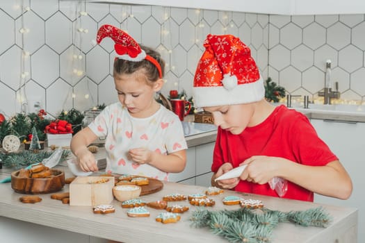 Little dark-haired girl 3 years old and boy 8 years old in red Santa hat decorate gingerbread cookies with icing. Siblings in white Christmas decorated kitchen. Merry Christmas, Happy New Year