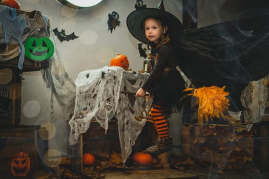 Little girl 3-4 years old in witch costume and striped stockings flies on broom in Halloween yard decoration with orange bright halloween pumpkins and wooden boxes with gauze as web, bats and spiders