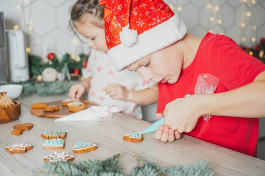 Little dark-haired girl 3 years old and boy 8 years old in red Santa hat decorate gingerbread cookies with icing. Siblings in white Christmas decorated kitchen. Merry Christmas, Happy New Year