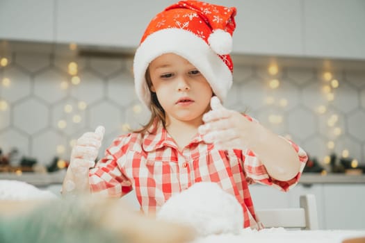 Little dark-haired girl 3 years old in red Christmas cap and checkered shirt prepares dough for gingerbread cookies in white Christmas decorated kitchen with garland lights. Merry Christmas, New Year